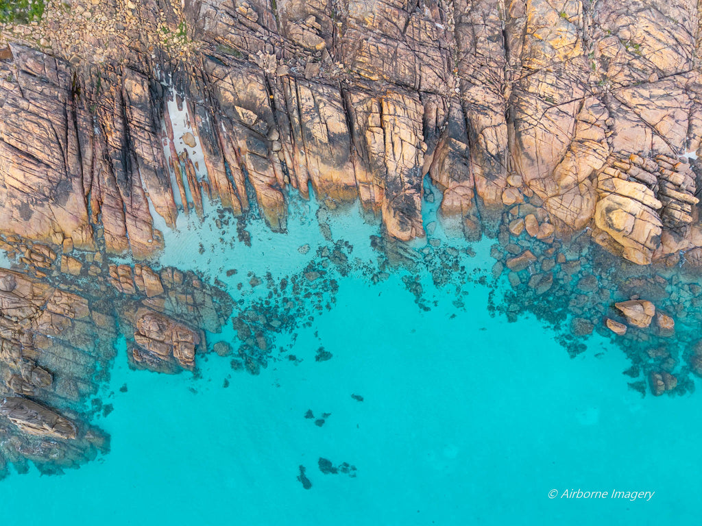 Overhead aerial view of Meelup Beach’s rocky coastline, with weathered granite ledges bordering vivid turquoise water and submerged boulders visible below.