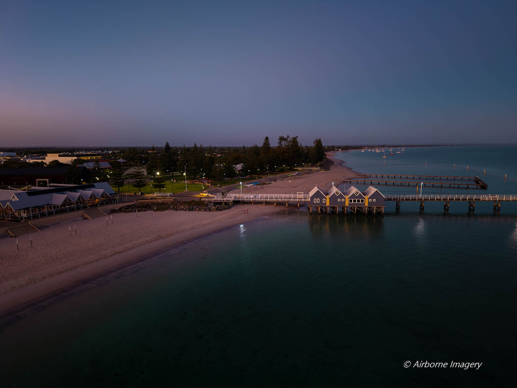 Aerial view of Busselton Jetty at dawn, with illuminated jetty buildings, a sandy beachfront and calm Geographe Bay beneath a blue and pink sky.