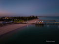 Aerial view of Busselton Jetty at dawn, with illuminated jetty buildings, a sandy beachfront and calm Geographe Bay beneath a blue and pink sky.