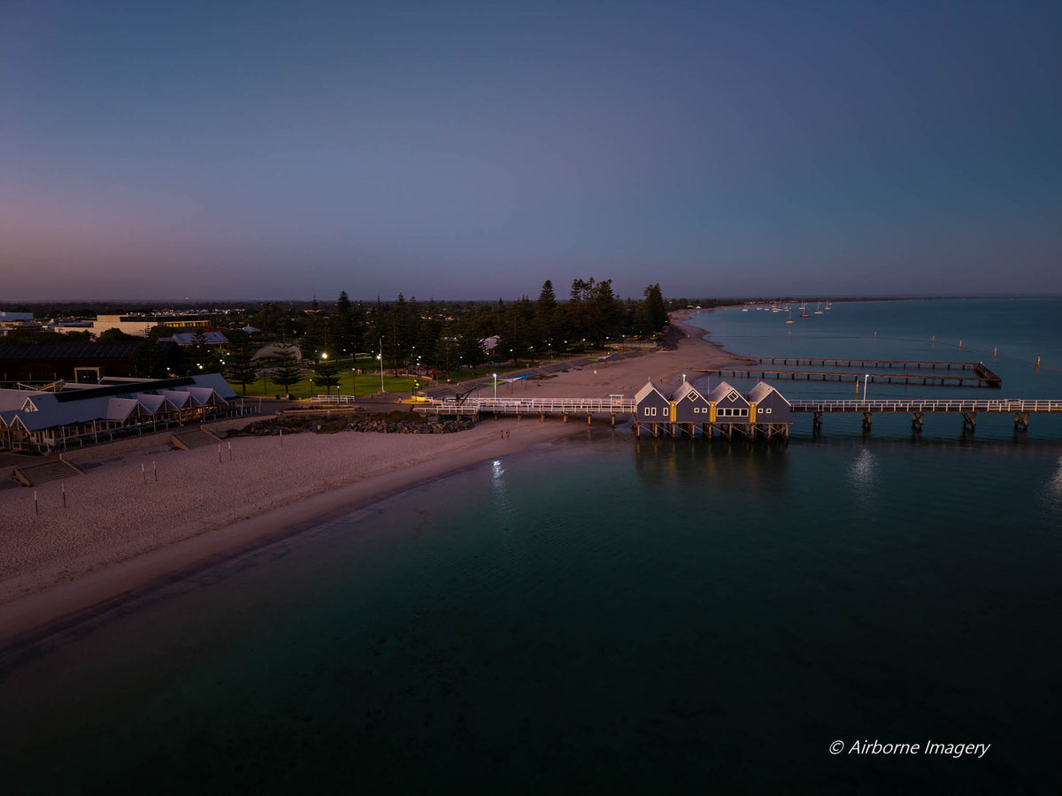 Aerial view of Busselton Jetty at dawn, with illuminated jetty buildings, a sandy beachfront and calm Geographe Bay beneath a blue and pink sky.