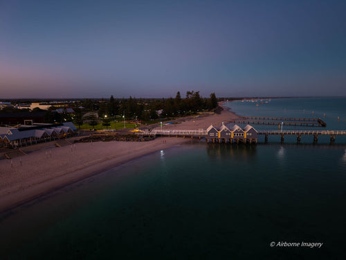 Aerial view of Busselton Jetty at dawn, with illuminated jetty buildings, a sandy beachfront and calm Geographe Bay beneath a blue and pink sky.