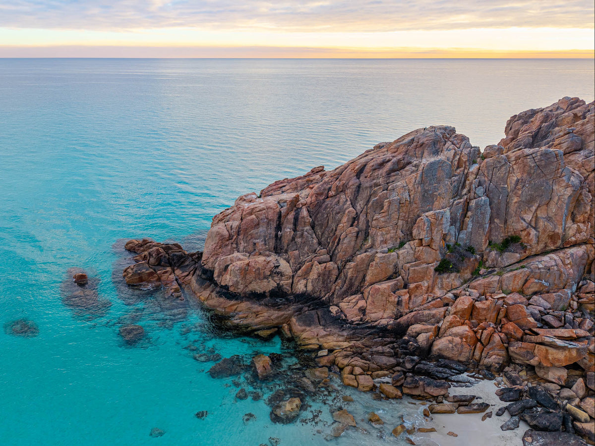 Aerial view of Castle Rock in Dunsborough, with rugged pink granite, clear turquoise water and a small sandy cove beneath a softly glowing golden sky.