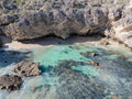 Aerial view of a secluded cove at Three Bears, with rugged cliffs, a narrow sandy beach and clear turquoise shallows revealing dark reef and scattered rocks.