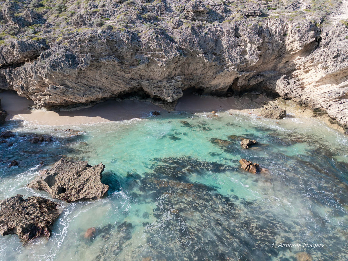 Aerial view of a secluded cove at Three Bears, with rugged cliffs, a narrow sandy beach and clear turquoise shallows revealing dark reef and scattered rocks.