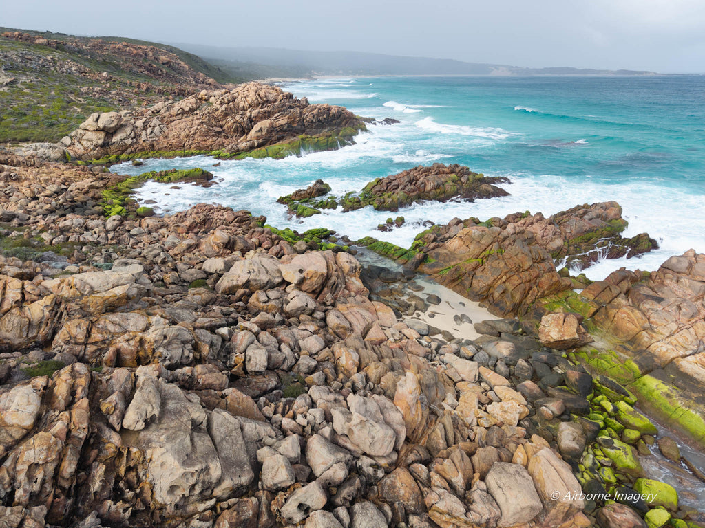 Aerial view of Conto’s Beach with rugged granite boulders, vivid green algae-covered rocks and white waves breaking along a turquoise coastline.