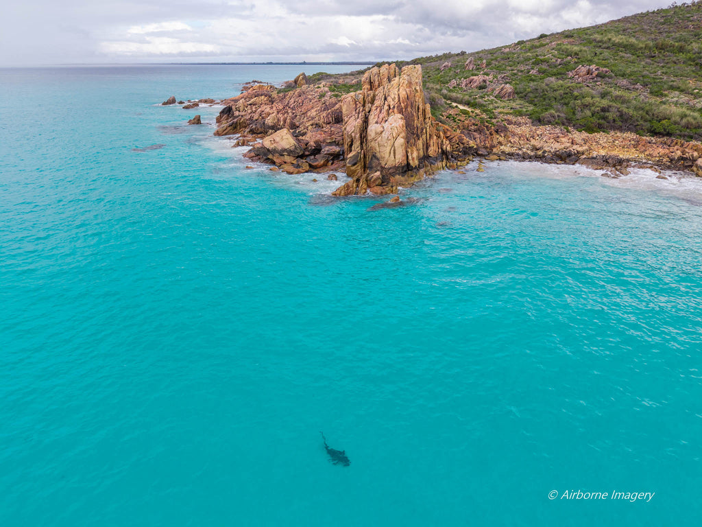 Aerial view of Castle Rock in Dunsborough, with clear turquoise shallows, a pink granite outcrop, a small sandy beach and coastal greenery beneath a cloudy sky.