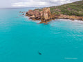 Aerial view of Castle Rock in Dunsborough, with clear turquoise shallows, a pink granite outcrop, a small sandy beach and coastal greenery beneath a cloudy sky.