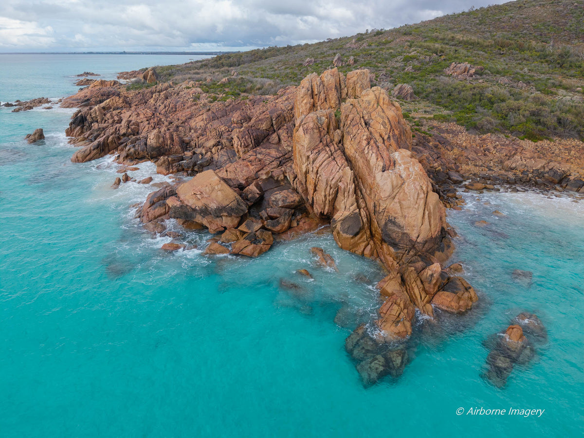 Aerial view of Castle Rock’s towering pink granite formation surrounded by turquoise water, with white surf, coastal scrub and a cloudy sky.