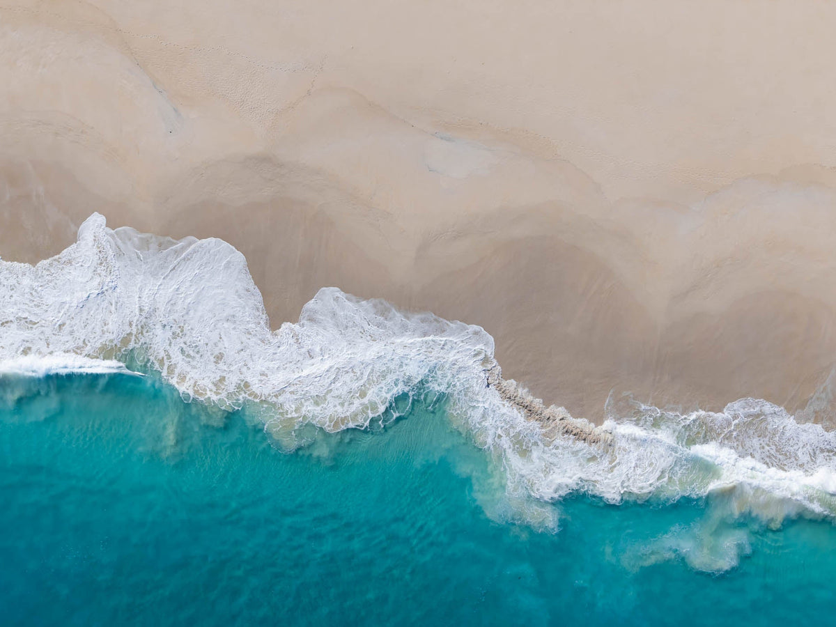 Overhead aerial view of Injidup Beach in Yallingup, with a foamy white wave sweeping diagonally between turquoise ocean and smooth pale sand.