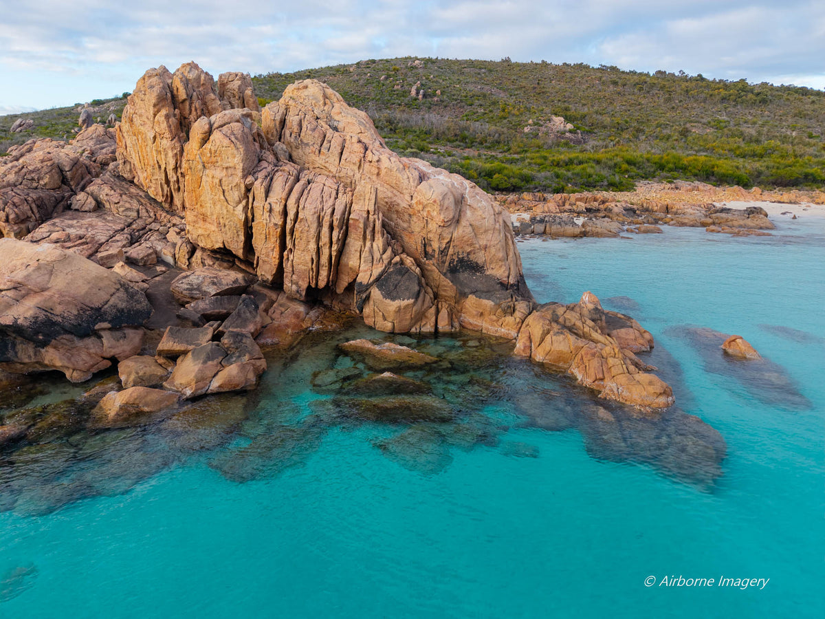 Aerial view of Castle Rock in Dunsborough, with sculpted pink granite rising from clear turquoise water and a scrub-covered hillside beneath a cloudy sky.