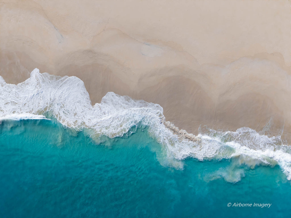 Overhead aerial view of Injidup Beach in Yallingup, with a foamy white wave sweeping diagonally between turquoise ocean and smooth pale sand.