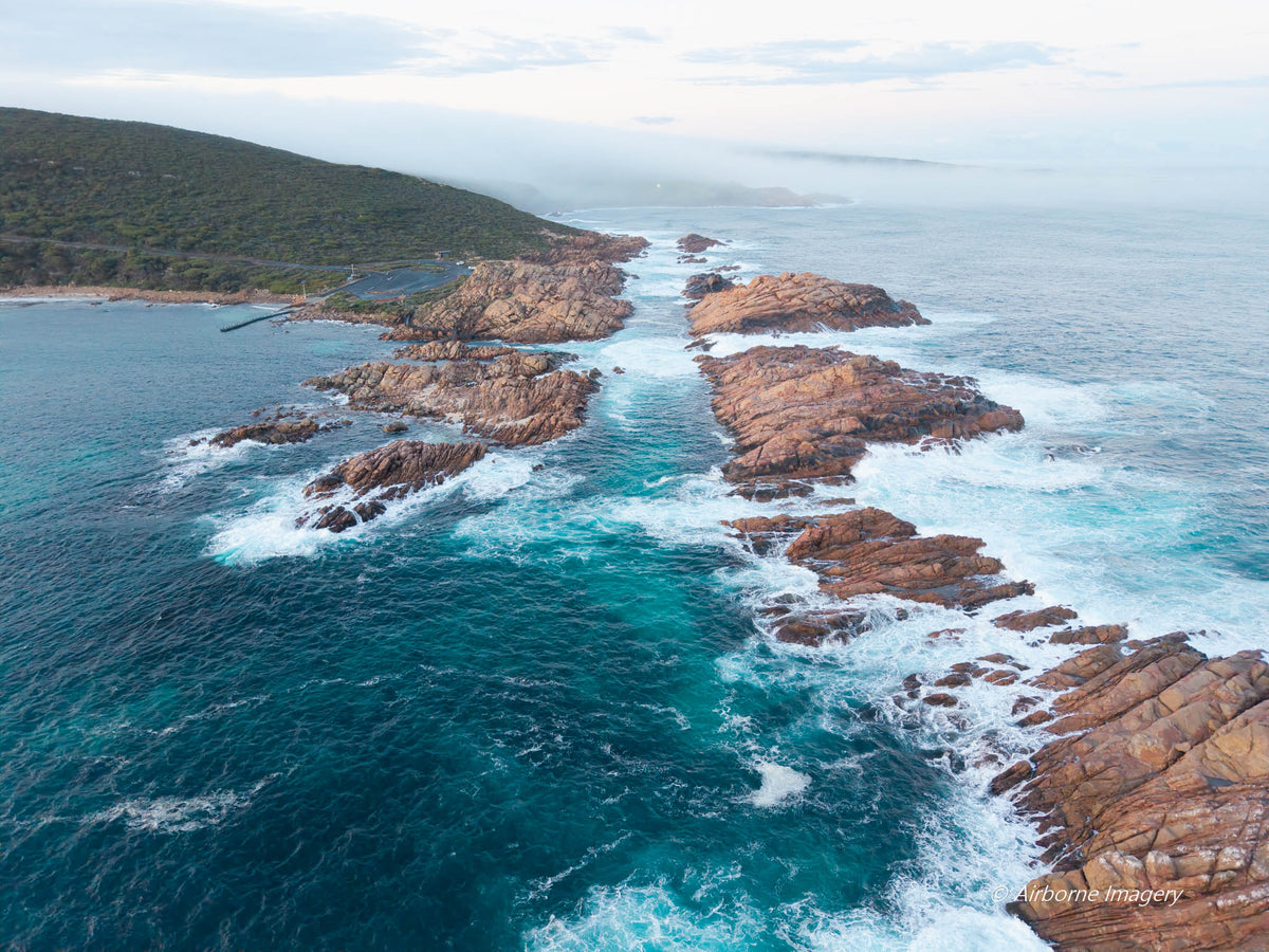 Aerial view of Canal Rocks in Yallingup, with deep blue water flowing between pink granite outcrops, breaking white surf and a misty coastal hillside.