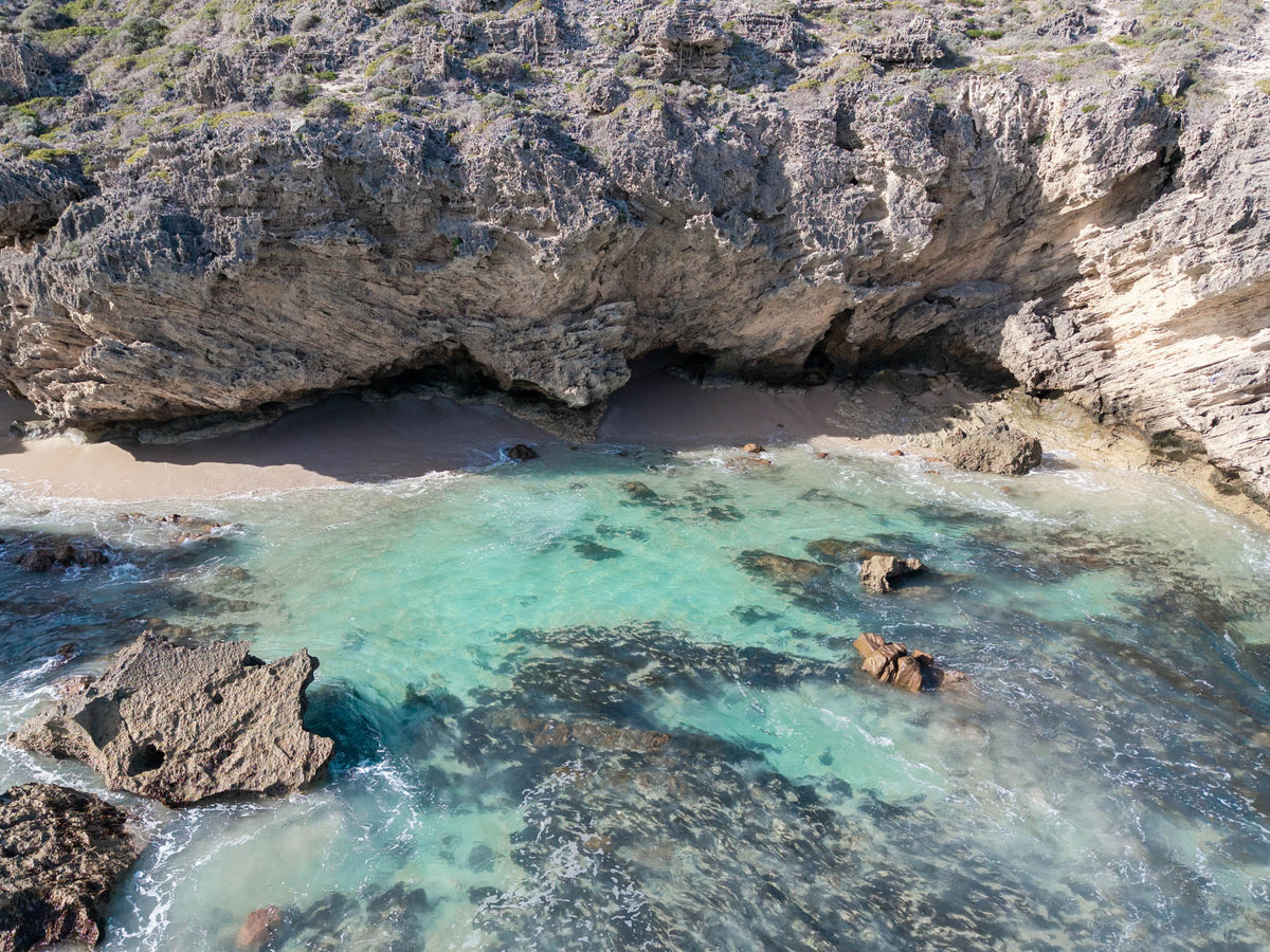 Aerial view of a secluded cove at Three Bears, with rugged cliffs, a narrow sandy beach and clear turquoise shallows revealing dark reef and scattered rocks.