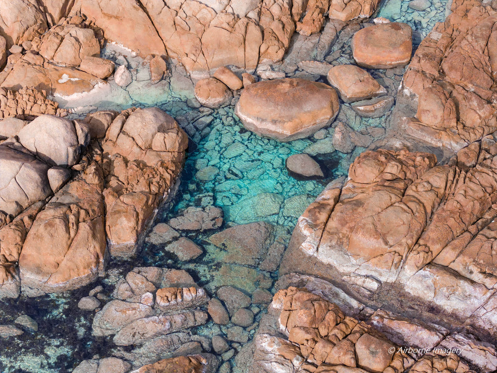 Overhead aerial view of Eagle Bay, with clear turquoise water between weathered pink granite boulders and submerged rocks visible beneath the surface.