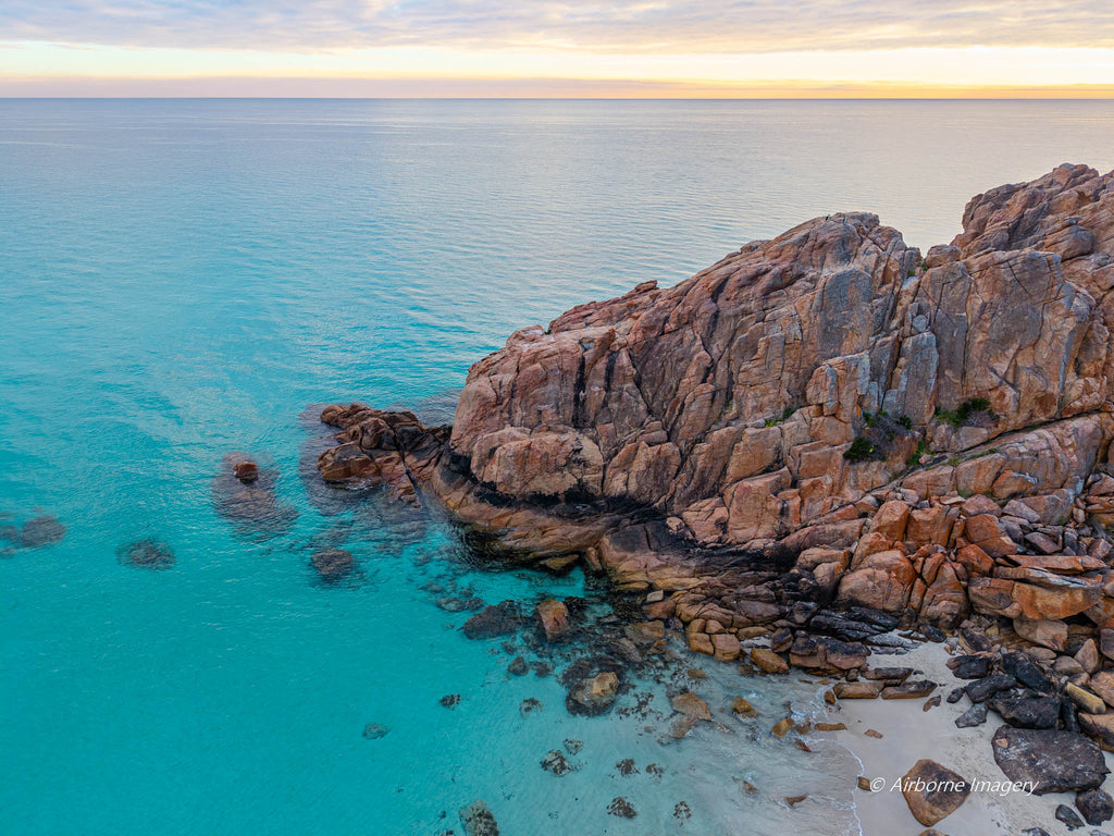 Aerial view of Castle Rock in Dunsborough, with rugged pink granite, clear turquoise water and a small sandy cove beneath a softly glowing golden sky.